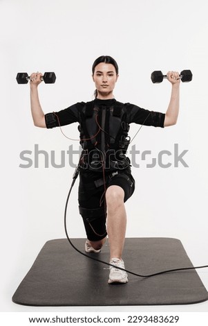 Fit girl with dumbbells in EMS suit that uses electrical impulses to stimulate muscles on white background. Sport training in electrical muscle stimulation suit