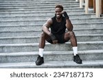 Fit happy sporty young black man boxer fighter sitting at urban stairs feeling confident. Strong African ethnic guy relaxing after street workout exercises or boxing, resting after training outside.