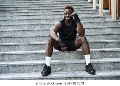 Fit happy sporty young black man boxer fighter sitting at urban stairs feeling confident. Strong African ethnic guy relaxing after street workout exercises or boxing, resting after training outside. - Powered by Shutterstock
