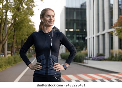 A fit and happy woman takes a break from running, her hands on her hips as she pauses to enjoy the city scenery. Earphones in, she's dressed in athletic gear, ready to continue her fitness routine. - Powered by Shutterstock