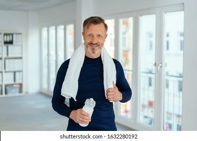 Fit Healthy Mature Man After A Workout At Work Standing Holding A Bottle Of Fresh Water And Towel Around His Neck Smiling At The Camera In A Corporate Gym