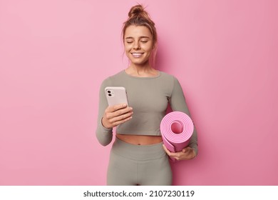 Fit healthy woman holds rolled fitness mat modern mobile phone downloads application for sport training dressed in activewear isolated over pink background. People and regular workout in gym