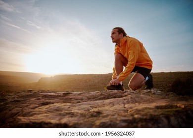 Fit male jogger tying shoelace preparing for cross country mountain trail in nature and looking away against sunrise
