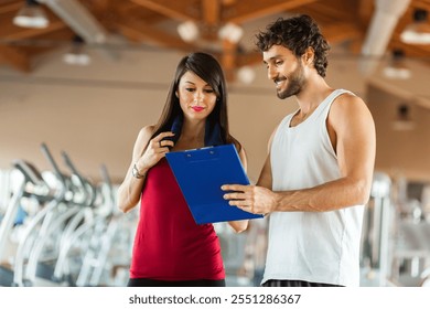 Fit male trainer discusses progress with a female client holding a clipboard in a gym setting - Powered by Shutterstock