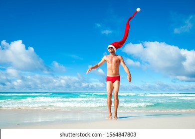 Fit man with extra long Santa hat celebrating the holidays in Christmas red swimming briefs on the shore of a tropical beach