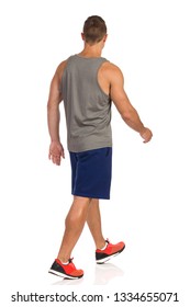 Fit Man In Gray Tank Top, Blue Shorts And Orange Sneakers Is Walking And Looking Away. Rear Side View. Full Length Studio Shot Isolated On White.