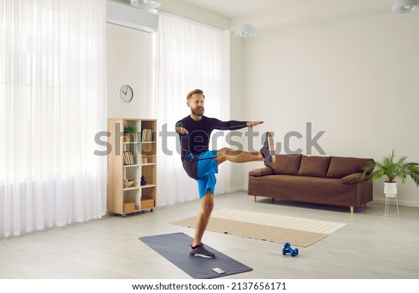 Fit man having active workout at home. Handsome\
strong athlete following individual fitness program doing leg swing\
exercise on sports mat and using modern mobile phone app to track\
physical activity