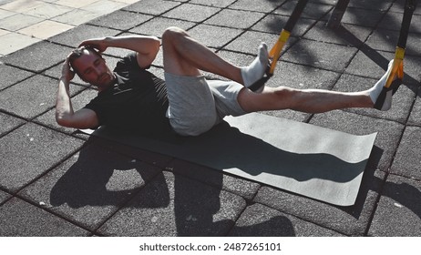 A fit man lays on a yoga mat outside at an outdoor gym and performs bicycle crunches to strengthen his core. He is dressed in fitness attire for a workout. - Powered by Shutterstock