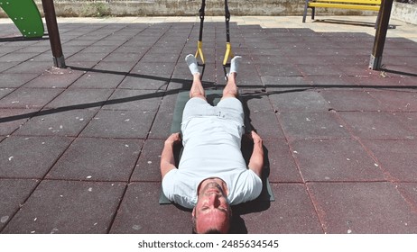 A fit man lies on an exercise mat outdoors on a sunny day. He is using fitness straps to perform a leg exercise. He is focused on his workout routine. - Powered by Shutterstock