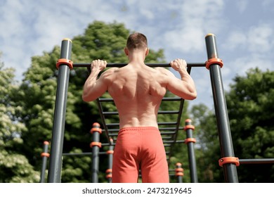 A fit man in red shorts exercises on outdoor gym equipment, doing pull-ups with his muscular back visible under the open sky, showcasing strength and fitness. - Powered by Shutterstock