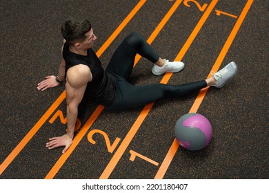 Fit Man Sitting On Gym Floor. Athlete Resting After Fitness Ball Workout In Gym. Fitness Training