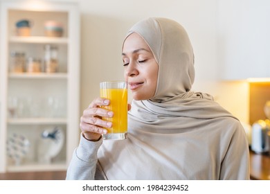 Fit mature arabian muslim islamiic woman in hijab drinking orange juice fresh smoozie in the kitchen, healthy eating breakfast, organic food
