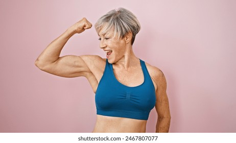 A fit, mature woman with short grey hair flexes her muscles against a pink isolated background, displaying strength and vitality. - Powered by Shutterstock