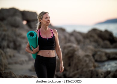 Fit middle age woman holding a sports mat and preparing to practice yoga on sea beach. - Powered by Shutterstock