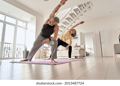 Fit middle aged 50s family couple doing fitness yoga morning exercise at home. Sporty healthy old mature man and woman training together standing in living room. Active seniors sport stretching.