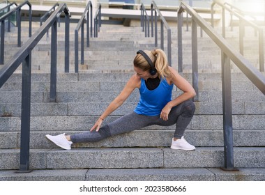 Fit Middle Aged Woman In Headphones Stretching Legs While Working Out Outdoors In The City