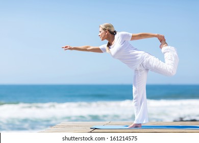 Fit Middle Aged Woman In White Doing Yoga Exercise At The Beach