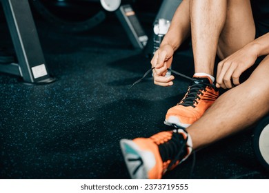 A fit and muscular male is tying his shoelace on the gym floor, surrounded by dumbbells and other workout equipment. The shot emphasizes his commitment to fitness and bodybuilding. - Powered by Shutterstock