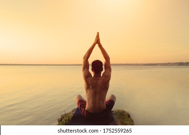 Fit Muscular Man Doing Yoga By The Water Sitting In A Lotus Pose With Hands Reaching Up The Sky Namaste. Orange Sunlight Sunset Tranquility At Lake. Male Back Defined Muscles. Healthy Spine Workout