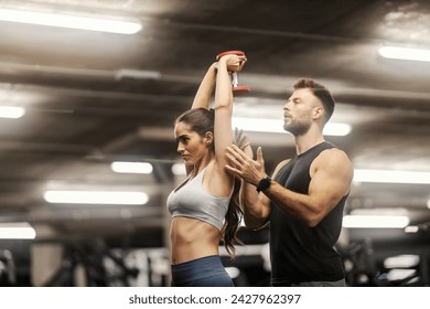 A fit muscular sportswoman in shape is doing triceps extensions with dumbbell in a gym with a trainer's little help.