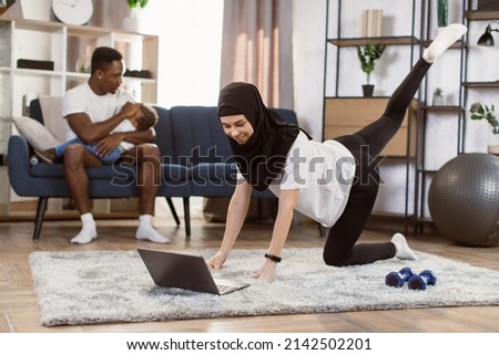 Fit muslim arabic woman doing yoga plank and watching online tutorials on laptop while training in living room and her african husband puts little son to sleep while sitting on the couch.