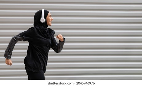 Fit muslim woman in modest sportswear and wireless headphones running outdoors, side view shot in motion of athletic islamic female runner in hijab doing sports outside, panorama with copy space