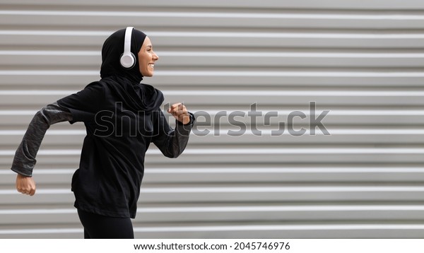 Fit muslim woman in modest sportswear and wireless\
headphones running outdoors, side view shot in motion of athletic\
islamic female runner in hijab doing sports outside, panorama with\
copy space