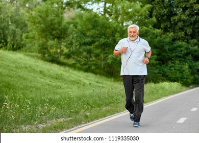 Fit old man running on racetrack in green park. Outdoors sport, trainings. Wearing classic white polo shirt with dark blue stripes,black trousers, sneakers. Listening to music with red headphones.