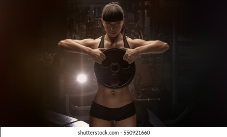 Fit Power Athletic Confident Young Woman Crossfit Trainer Doing Exercises With Heavy Weight Barbell Plate In Gym Rising Hand. Fitness Muscular Body, Strong Hand On Dark Background Pumping Up Muscles