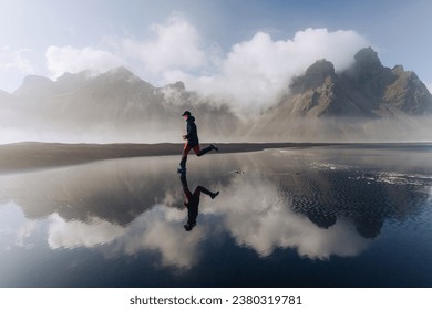 A fit runner is running on the seashore in the wild nature of Iceland. An explorer is exercising in nature in Iceland. There are mountains in the background.