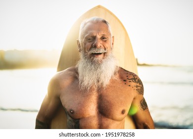 Fit Senior Male Having Fun Surfing During Sunset Time - Retired Man Training With Surfboard On The Beach - Elderly Healthy People Lifestyle And Extreme Sport Concept