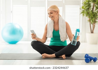 Fit senior woman relaxing on yoga mat with smartphone after her home workout, checking sports videos online or browsing social networks, beautiful mature lady taking break after yoga practice - Powered by Shutterstock