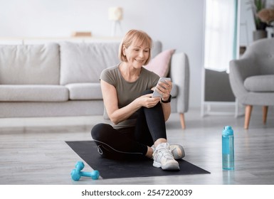 Fit senior woman relaxing on yoga mat with smartphone after her home workout, checking new sports videos online. Mature Caucasian lady taking break after yoga practice. Active lifestyle concept - Powered by Shutterstock