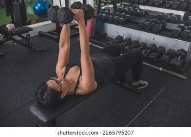 A fit short haired asian woman does a set of close grip dumbbell bench presses, also known as crush press. Working out and lying on a flat bench at the gym. Training chest and triceps. - Shutterstock ID 2265795207