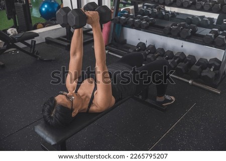 A fit short haired asian woman does a set of close grip dumbbell bench presses, also known as crush press. Working out and lying on a flat bench at the gym. Training chest and triceps.