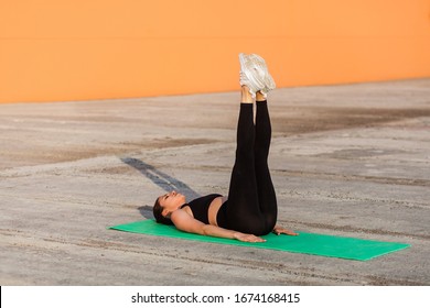 Fit slim young woman in tight sportswear, black pants and top, lying on mat keeping straight legs raised, training abdominal muscles, flexibility. Health care and weight loss, sport activity outdoor - Powered by Shutterstock