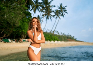 Fit solo female traveler enjoys tropical beach getaway, slender woman in bikini relaxes by sea amidst palm trees, exemplifies wellness travel. - Powered by Shutterstock