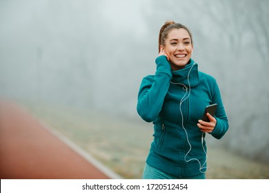 Fit Sportswoman With Beautiful Smile Jogging Outside On Cold And Foggy Morning. The Girl Is Listening To Music. 