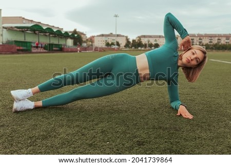 Fit sportswoman in blue activewear doing side plank exercise while balancing on green field at stadium during workout in summer and looking at camera 