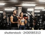 A fit sportswoman in shape is doing exercises with dumbbells in a gym and her coach is helping her.