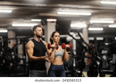 A fit sportswoman in shape is doing exercises with dumbbells in a gym and her coach is helping her. - Powered by Shutterstock