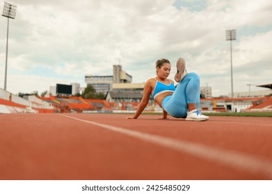 A fit sportswoman is sitting on running track at stadium, stretching for exercising. A fit runner in shape is sitting at stadium and doing warm up exercises. A sporty woman stretching legs outdoors. - Powered by Shutterstock