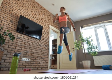 Fit Sporty Active Young Adult Woman Wearing Sportswear Doing Fitness Sport Exercise At Home. Athletic Strong Girl Jumping Up In Air Indoor In Modern Apartment Living Room. At Home Gym Workout Concept.