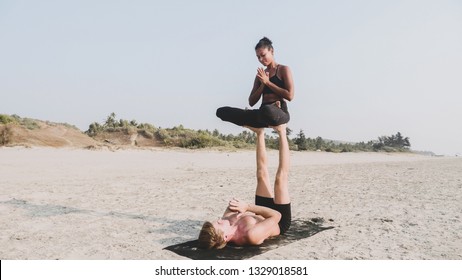 Fit sporty couple practicing acro yoga with partner together on the sandy beach. Female acrobat sits in lotus pose on the feet of her male partner. Mix race couple doing acrobatic exercise