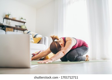 Fit Sporty Healthy Woman On Mat In Child’s Balasana Gentle Resting Yoga Pose, Doing Breathing Exercises, Watching Online Yoga Class On Laptop Computer. Healthy People And Self Motivation Concept.