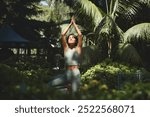 Fit sporty mindful young Hispanic woman meditating doing yoga breathing exercises with eyes closed feeling peace of mind, mental balance standing in green nature tropical park on sunny day.