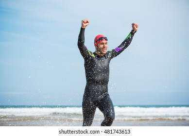 Fit Sporty Open Water Swimmer Man Running Off Shore On A Beach Rising Arms Up In Victory Sign After Swimming Triathlon Competition Exercise Routine Workout.