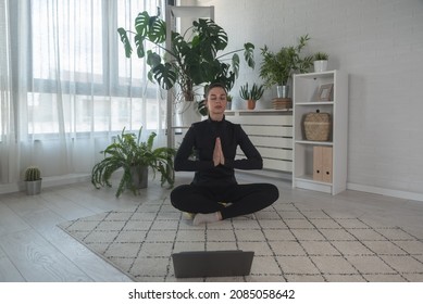 Fit sporty woman doing yoga Sukhasana easy sitting pose in living room at home watching online tutorials from trainer on laptop. Workout, fitness sport and healthy lifestyle concept with meditation.