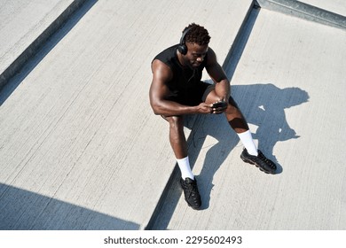 Fit sporty young black man sitting on concrete urban stairs holding phone using mobile apps listening music. Strong African ethnic guy wearing headphones looking at smartphone outdoors. Top view - Powered by Shutterstock