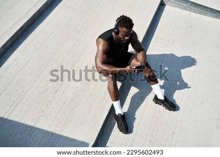 Fit sporty young black man sitting on concrete urban stairs holding phone using mobile apps listening music. Strong African ethnic guy wearing headphones looking at smartphone outdoors. Top view